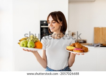 Choosing between healthy or unhealthy food. Young woman holding plates with fruits and sweets, looking at healthy snack with smile, deciding what to eat, standing in kitchen interior Royalty-Free Stock Photo #2057026688
