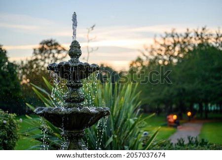 Fountain with water drops in a park with trees behind at dusk. The fountain is seen with a path, bench and lights just after sunset. This fountain is a central feature in the park. Royalty-Free Stock Photo #2057037674