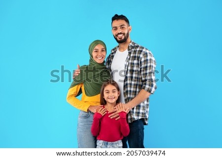 Modern Muslim Family Of Three Hugging Smiling To Camera Standing Together Over Blue Studio Background. Islamic Mother In Hijab And Father Embracing Their Happy Little Daughter