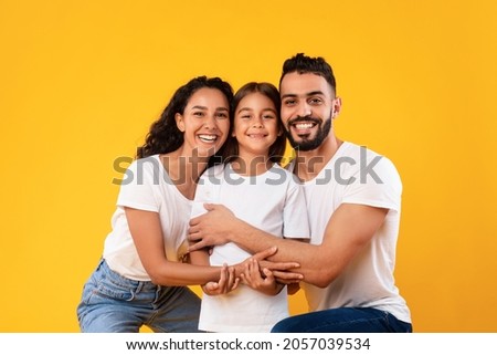 Arab Family. Happy Middle-Eastern Parents Hugging Their Kid Daughter Smiling To Camera On Yellow Background. Studio Shot Of Spouses Embracing Little Girl Expressing Their Love To Child