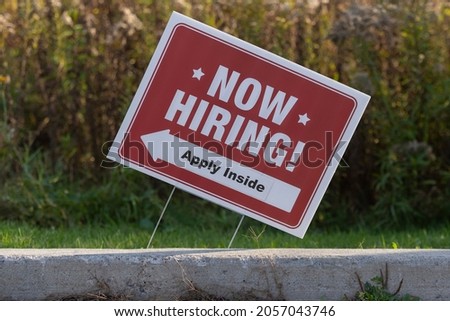 Outdoor lawn sign now hiring apply inside with direction arrow, selective focus. Great resignation, employment, understaffed business, work strikes, absent workers due to covid labor shortage concept.