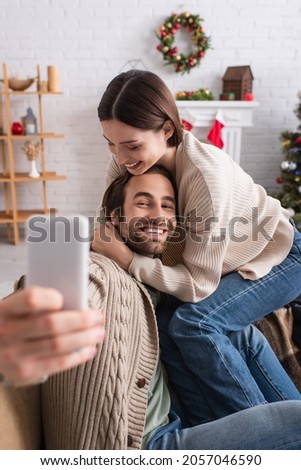 cheerful woman hugging husband taking selfie on blurred smartphone in decorated living room