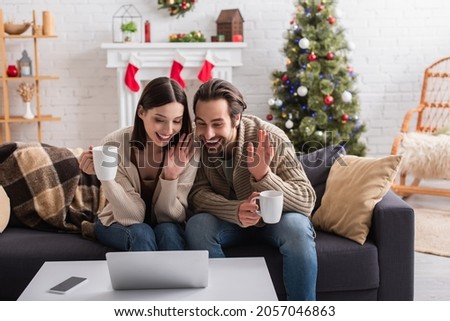 cheerful couple with tea cups waving hands during video call in decorated living room