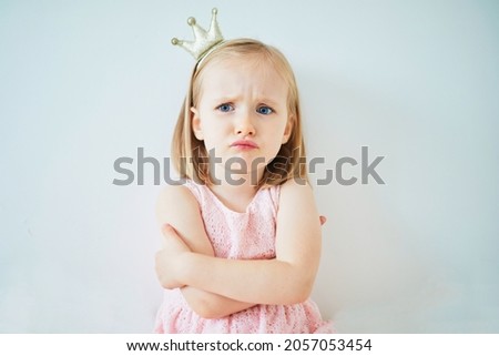 Adorable little girl in pink dress and golden crown dressed as princess. Child's portrait on light background. Child feeling angry, sad or gloomy