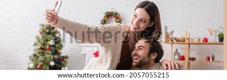 cheerful couple taking selfie on cellphone near blurred christmas tree in living room, banner