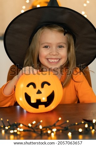 Little girl in witch costume and black hat looks at camera and smiles. Happy child celebrats Halloween with lantern pumpkin