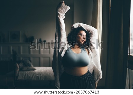 Curly haired plus size woman wearing comfortable clothes dances in stylish room.  Royalty-Free Stock Photo #2057067413