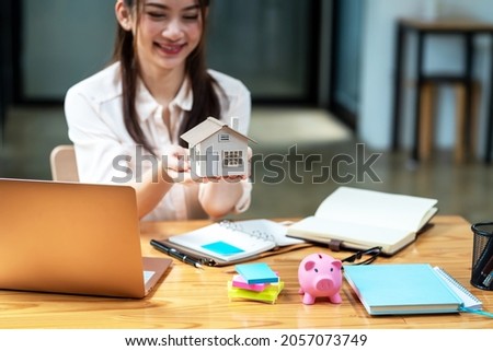 Young Asian businesswoman real estate agent holding house model with documents on table blurry background.