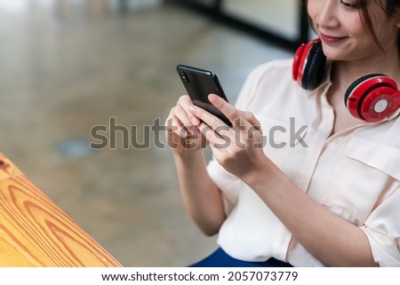 Image of a smiling young Asian woman wearing headphones holding a smartphone sitting in an office chair.