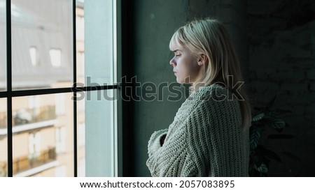 Beautiful Young Blonde Caucasian Woman Standing by the Window with Her Arms Crossed, Waiting for Someone To Visit Anxiously. Sad Girl Feeling Depressed and Hoping for Help.