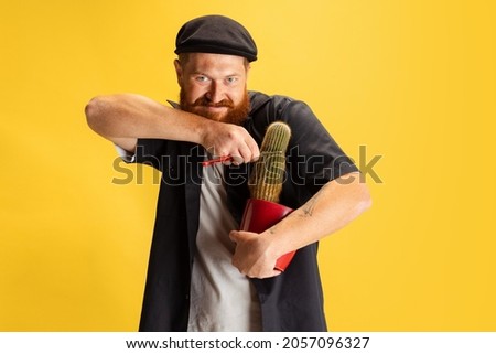 Cutting cactus. Comic portrait of stylish red-bearded man, barber in black cap having fun isolated over yellow background. Concept of emotions, facial expressions, fashion, style, funny meme emotions,