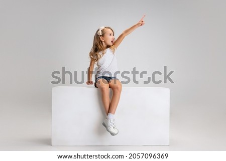 Pointing at. Portrait of little cute girl in casual clothes sitting on big box isolated on white studio background. Happy, joyful childhood, kids fashion, emotions, facial expression concept