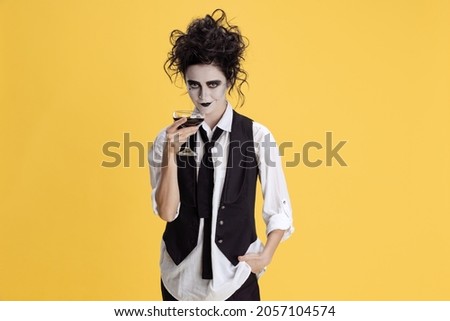 Cheers. Costume party gathering. Woman in black and white suit with creepy hair and make up drinking isolated on yellow background. Concept of party, costume, creativity, Halloween. Copy space for ad