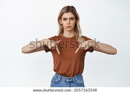 Angry and displeased young woman pointing fingers down, frowning upset, showing something disappointing, white background