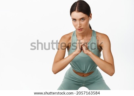 Fit fitness woman doing squats workout in gym, looking serious at camera, standing in activewear over white background