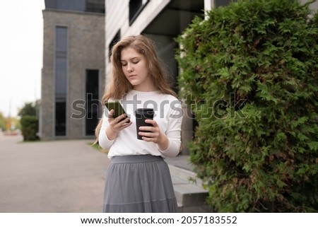 Successful female banker using smartphone outdoors while standing near office background, with paper cup of coffee. Young woman professional manager working on mobile device.