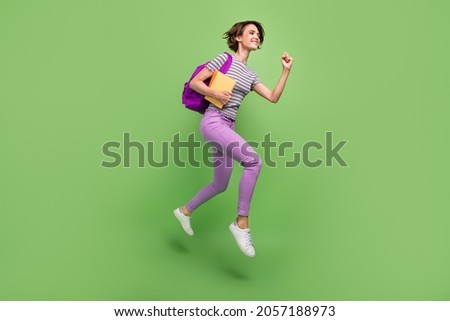 Photo of pretty hurrying young woman dressed striped t-shirt backpack copybooks jumping running smiling isolated green color background