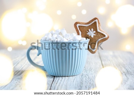 A blue cup of winter hot drink with marshmallows and gingerbread star on a white wooden background. Traditional Christmas atmosphere with bokeh. New year holiday concept. Festive background
