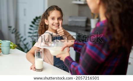 Closeup image of a mom pours her cute smiling daughter milk for breakfast, looking at camera shows thumb.