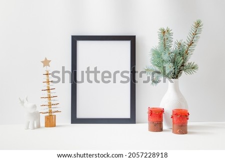 Christmas home interior with decor elements. Mockup with a black frame, spruce branches in a vase, candle, cone on a light background