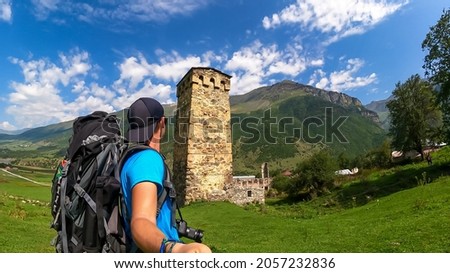 A man taking a picture wirh a lookout tower on a lush green pasture in Caucasus, Georgia. There are high mountain chains around. Clear and bright day with few clouds. Defense building. Hiking remedy