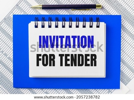 Against the background of reports on the desktop, a blue notepad. It has a pen and a white notebook with the text INVITATION FOR TENDER. Business concept