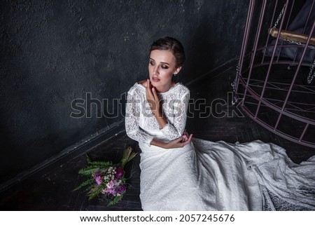 Fashion bride in a white openwork dress without a veil sitting by an iron cage, bars. Unhappy young woman near black textured background. Fictitious marriage. Wedding of convenience. Copy space Royalty-Free Stock Photo #2057245676