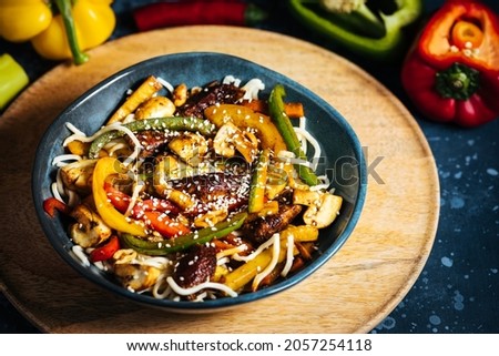 Wok with noodles, peppers, mushrooms, turkey meat and different vegetables and sesame seeds in a blue bowl on dark background