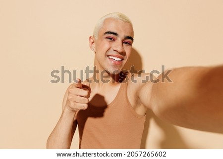Close up young latin gay man with make up in beige tank shirt do selfie shot pov on mobile phone point finger camera you isolated on plain light ocher background studio People lgbt lifestyle concept