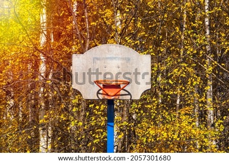 A red basketball hoop with a wooden battered shield without a net on the background of autumn yellow foliage of trees in the park.