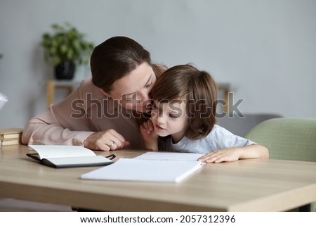 Cheerful young mother assisting her adorable son with schoolwork and exercises. Difficulties with school preparation and learning to write and draw. Happiness and support in the family