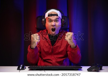 Excited and shocked face of Asian gamer with headphone playing computer pc video game online sitting at living room. Indian professional gamer streaming on social playing game very fun