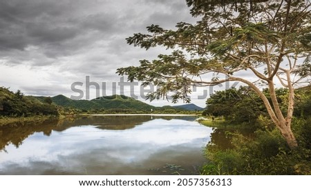Scenic Lake with Natural Background of Trees and Hills in Thailand. Cloudy Overcast Weather with Beautiful Reflections on Water Surface.