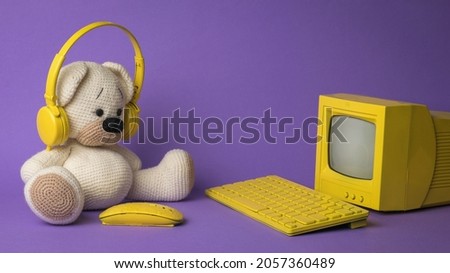 A knitted bear in front of a yellow computer on a purple background. The concept of virtual dependence of children.