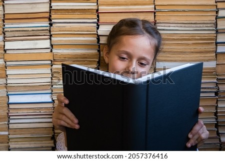 Curious cute preschool kid girl sitting with a book, on books background. Children reading addiction concept. Copy space on the book