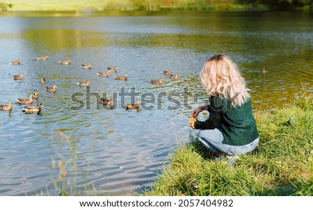 Side view of young woman in casual clothes sitting on shore near water and feeding ducks in nature. Selective focus on blonde girl throwing food to wild birds floating on lake.