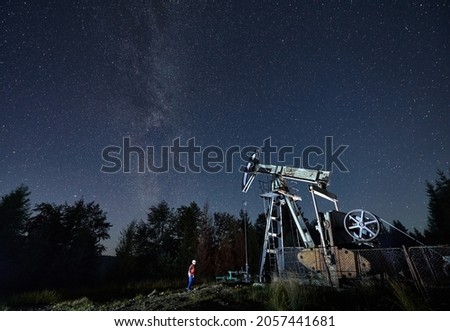 Beautiful view of night starry sky over oil field with petroleum pump jack and male worker. Oil man standing near oil pump rocker-machine under magical night sky with stars.