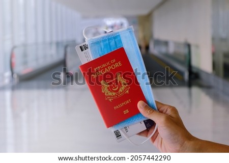 Travel concept: Hand holding SINGAPORE passport, with face mask and boarding pass in airport. Moving walkway travelator in background. Reopening travel lane for safe travels; covid-19 coronavirus. Royalty-Free Stock Photo #2057442290
