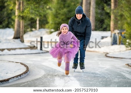 little girl in a pink sweater and a fluffy skirt on sunny winter day rides with her dad on an open skating rink in park