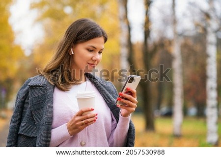 A beautiful girl stands in the park in autumn, holds a paper cup with coffee and looks into the phone Royalty-Free Stock Photo #2057530988