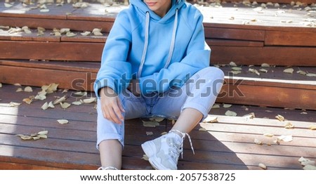 A girl is sitting on a park bench in a blue oversized hoodie.fashion and wear concept. warm oversize wear at female.space for text and logo.close up details of oversize wear.mock up.sneakers at model