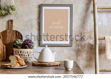 Stylish interior of kitchen space with wooden table, brown mock up photo frame, herbs, vegetables, tea pot, cups and kitchen accessories in wabi sabi concept of home decor. 