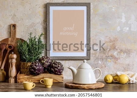 Stylish interior of kitchen space with wooden table, brown mock up photo frame, herbs, vegetables, tea pot, cup and kitchen accessories in wabi sabi concept of home decor. 