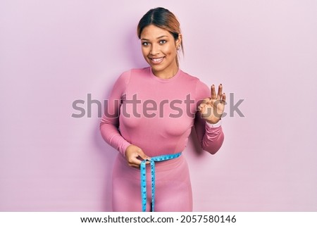 Beautiful hispanic woman using tape measure measuring waist doing ok sign with fingers, smiling friendly gesturing excellent symbol 