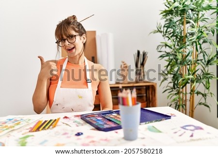 Hispanic woman at art studio smiling happy and positive, thumb up doing excellent and approval sign 