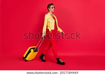 Photo of charming cool young woman dressed leather jacket dark glasses holding luggage walking smiling isolated red color background