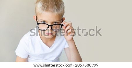 A blond-haired smart boy in black nerd glasses looks seriously at the camera with his glasses raised, a light beige monochrome background. Student, study, rigor concept. Copy space