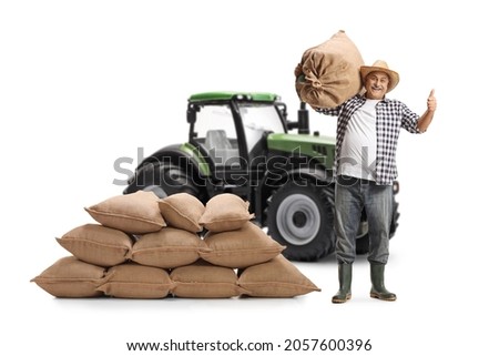 Full length portrait of a mature farmer with a pile of sacks gesturing thumbs up in front of a green tractor isolated on white background