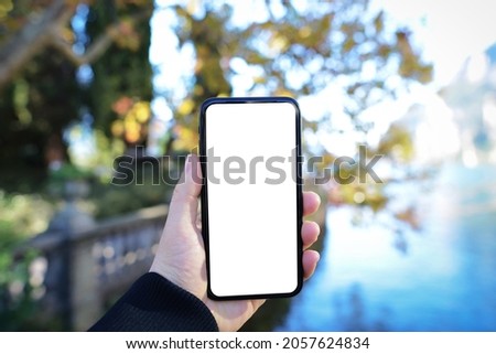Hands holding white screen smartphone. Mockup image of hand holding mobile phone with blank screen with green and lake nature background.