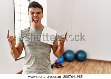 Young hispanic man wearing sportswear and towel at the gym shouting with crazy expression doing rock symbol with hands up. music star. heavy concept. 
